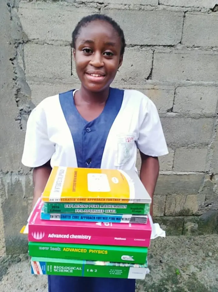 A secondary school student in uniform smiling and holding a tall stack of Advanced Level textbooks